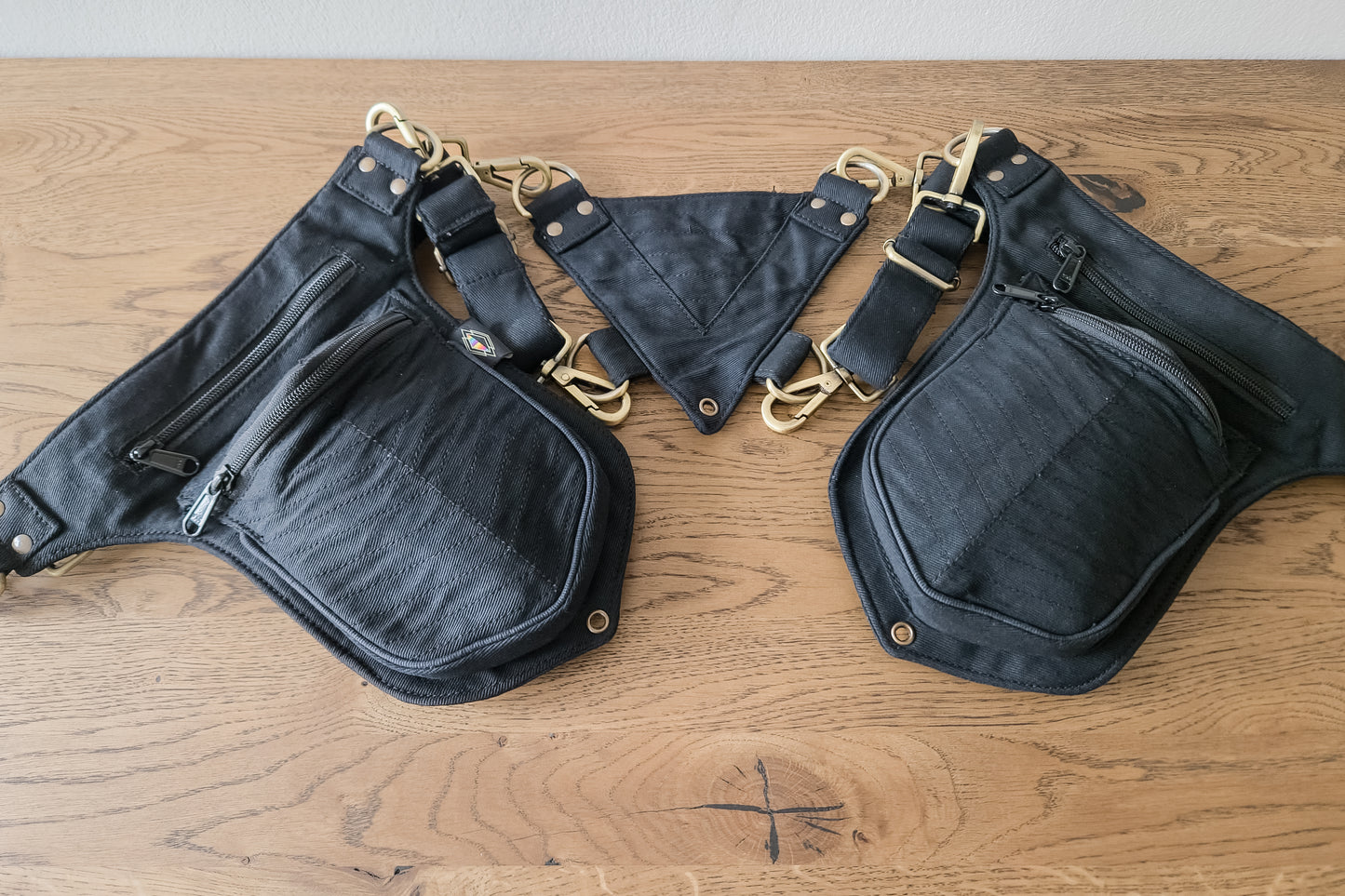 Schwarze Doppel-Schultertasche aus Baumwolle mit messingfarbenen Details und mehreren Reißverschlussfächern, flach auf Holztisch liegend.