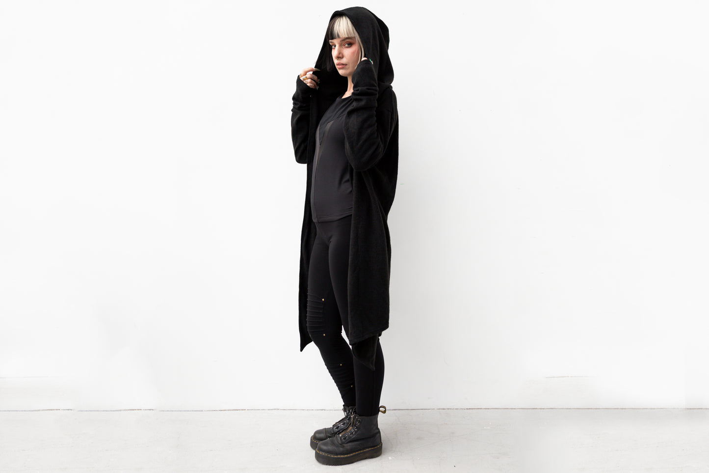 Model trägt langen schwarzen Strickmantel mit Kapuze im Goth-Stil, genderneutraler Schnitt, kombiniert mit schwarzen Leggings und Boots.
