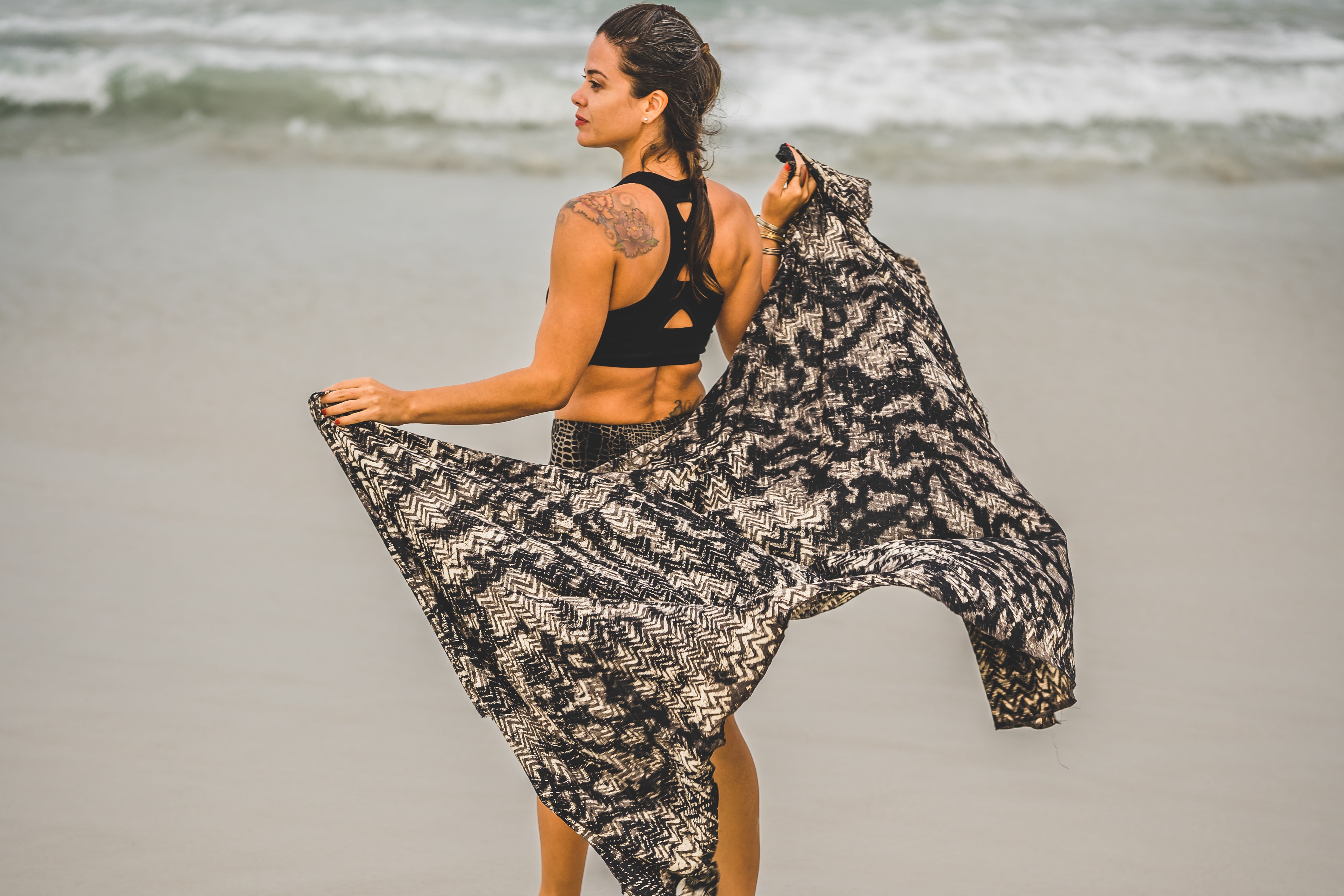 Model von hinten am Strand trägt einen schwarzen  Viskose-Sarong mit abstraktem Wellen-Batikmuster in Grau-Beige, vom Wind bewegt