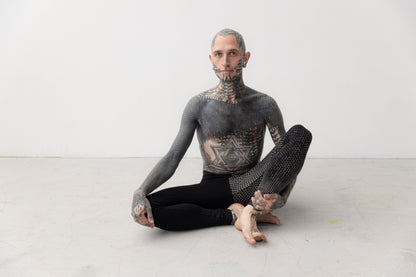 Männlich gelesenes Model trägt ausgefallene Leggings mit künstlerisch ungleichmäßigem Druck an einem Bein. Aufnahme im Sitzen.