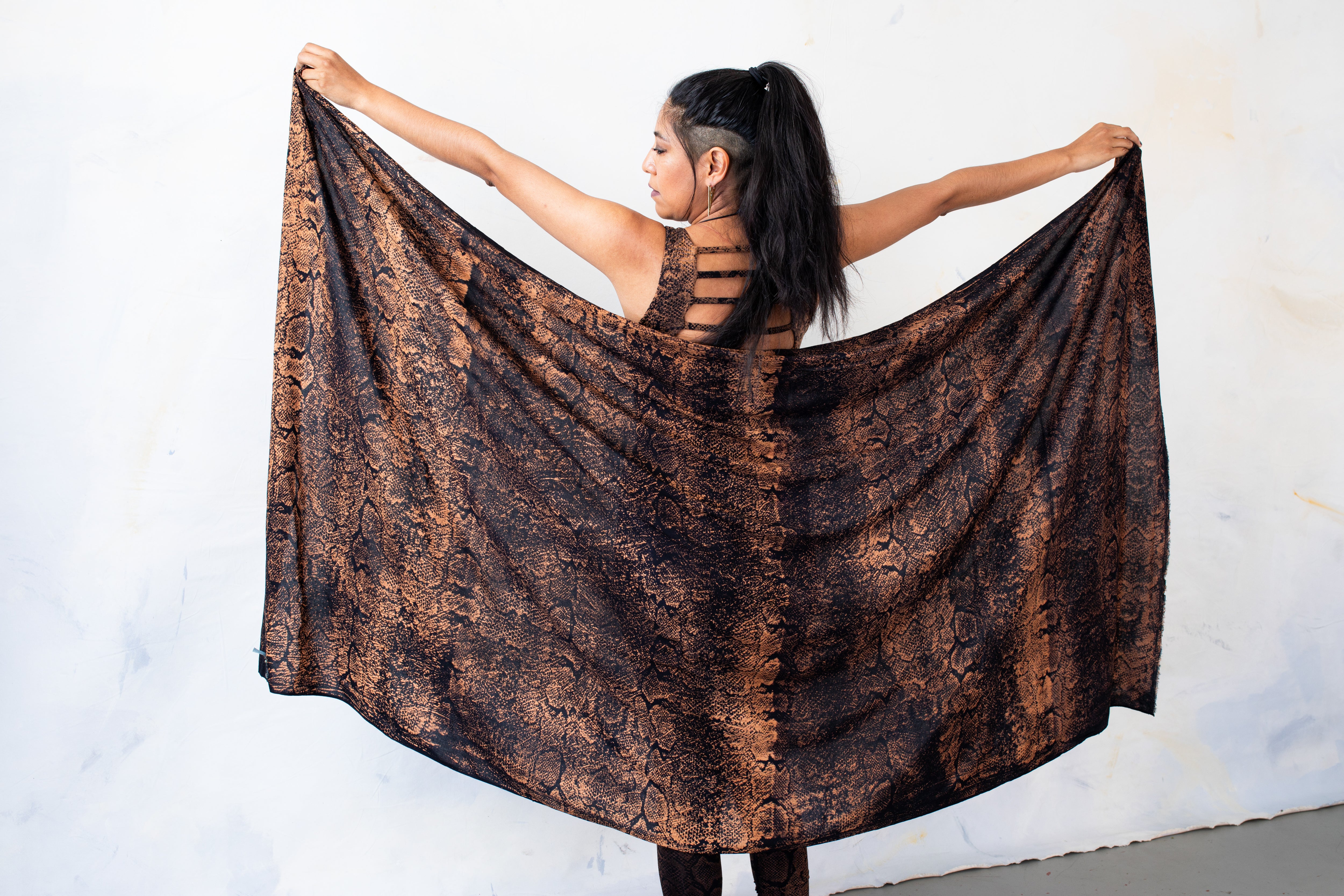 Model von hinten, das einen großen schwarzen Batik Sarong aus Viskose mit apricot-orangem Schlangenmuster mit beiden Armen aufspannt