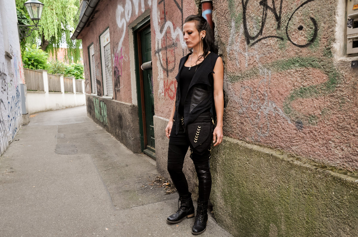 Model lehnt an Wand, trägt schwarze Bauchtasche mit Beinschnalle zu Kunstleder-Weste, seitliche Ganzkörperansicht