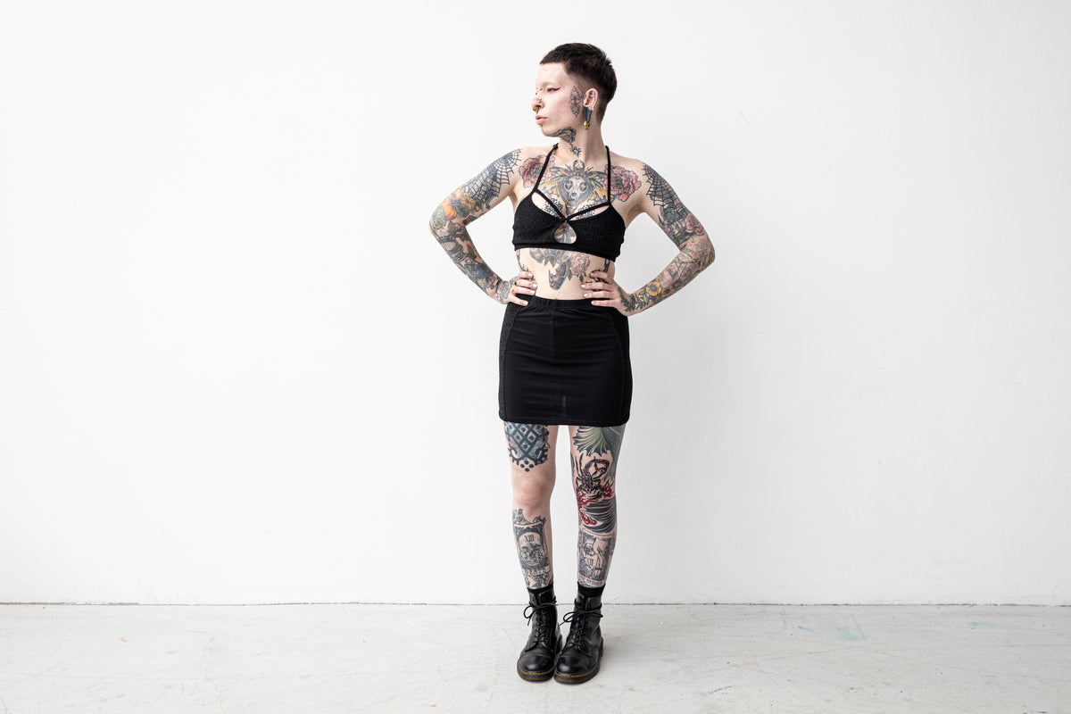 Weiblich gelesenes, alternatives Model trägt schwarzes Bralette aus gemusterter Spitze mit Riemchen-Detail.