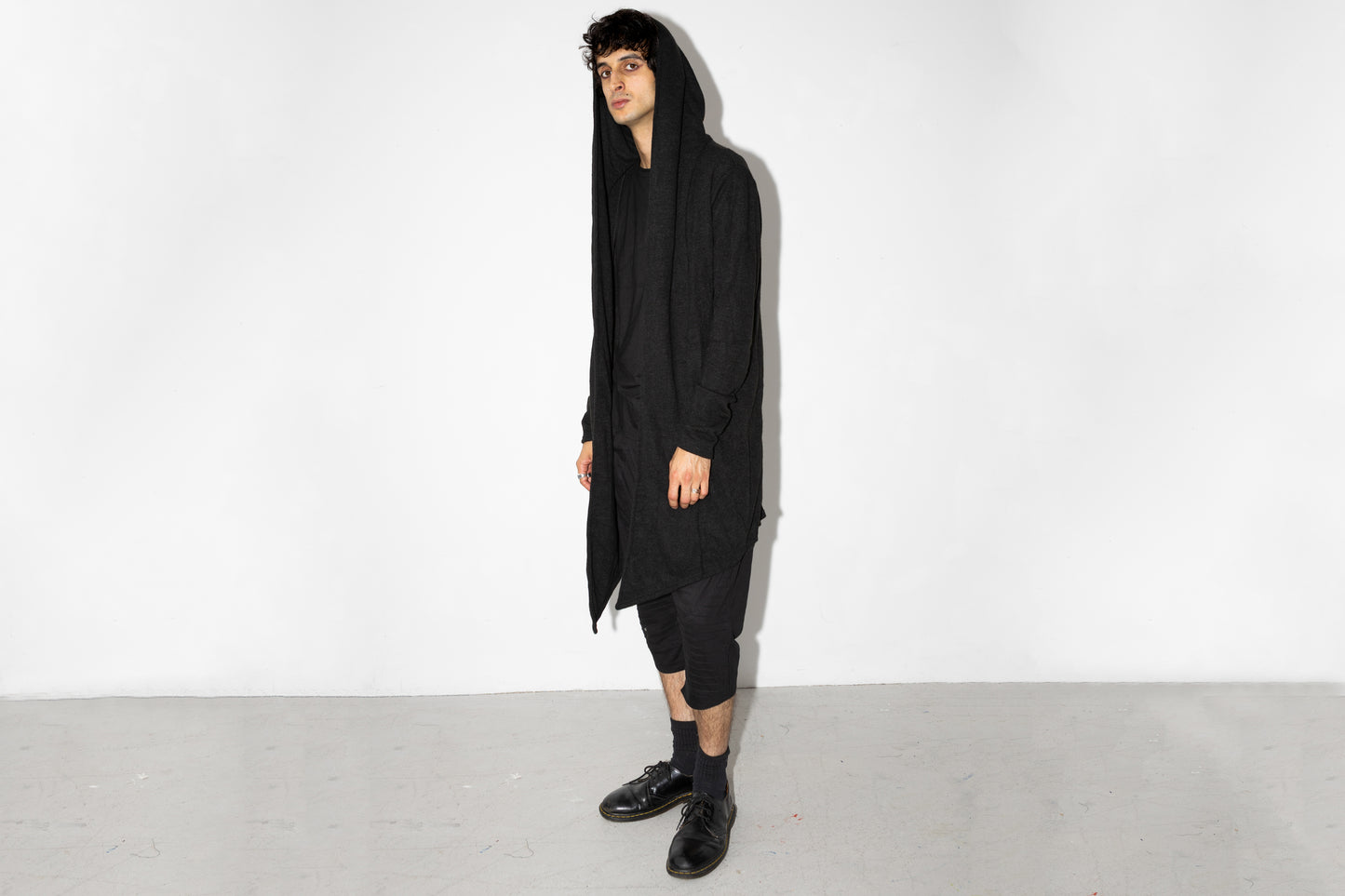 Model trägt langen schwarzen Strickmantel mit Kapuze im Goth-Stil, genderneutraler Schnitt, kombiniert mit schwarzer Hose und Halbschuhen.