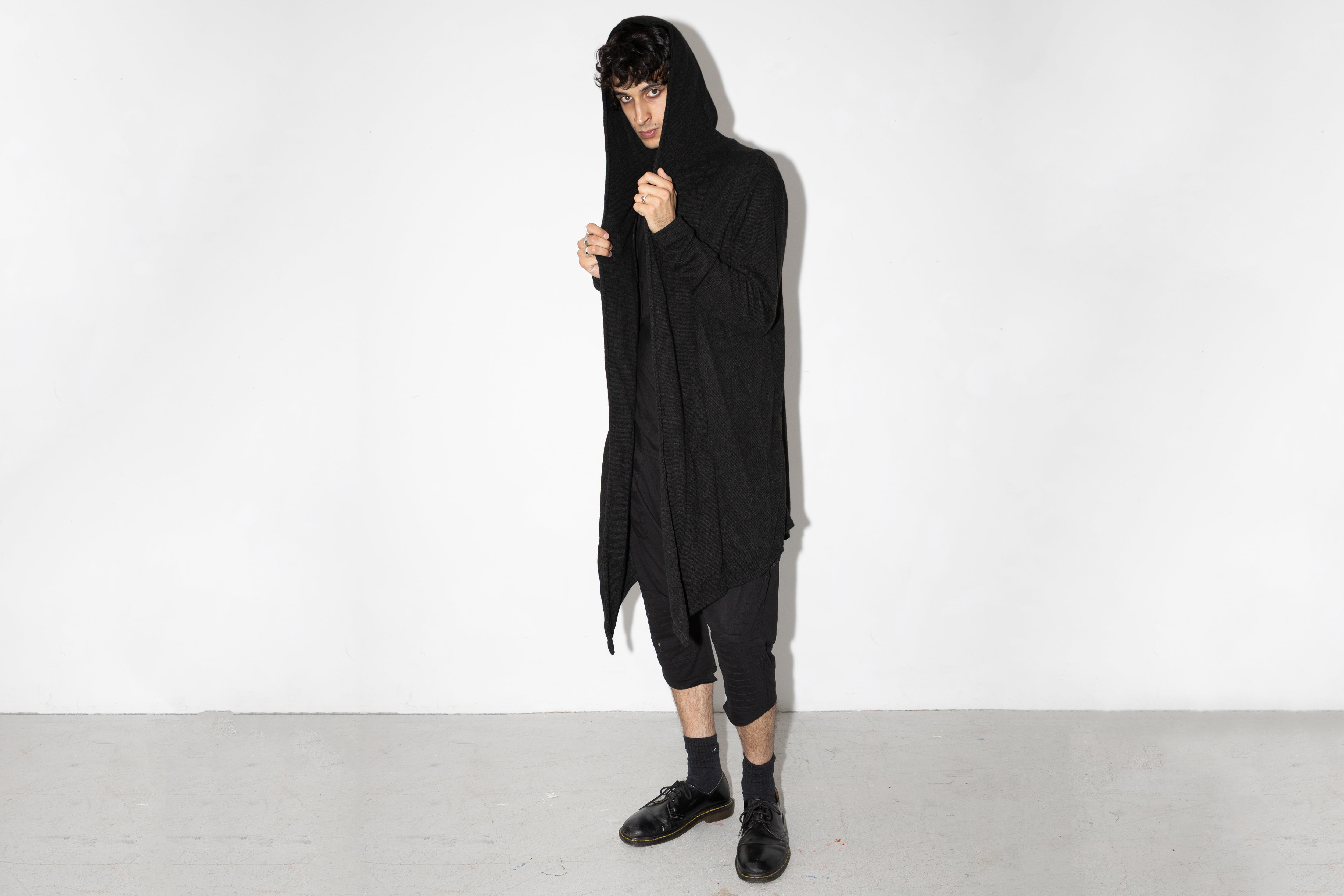 Model trägt langen schwarzen Strickmantel mit Kapuze im Darkwear-Stil, genderneutraler Schnitt, kombiniert mit schwarzer Hose und Halbschuhen.