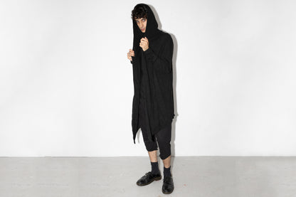 Model trägt langen schwarzen Strickmantel mit Kapuze im Darkwear-Stil, genderneutraler Schnitt, kombiniert mit schwarzer Hose und Halbschuhen.