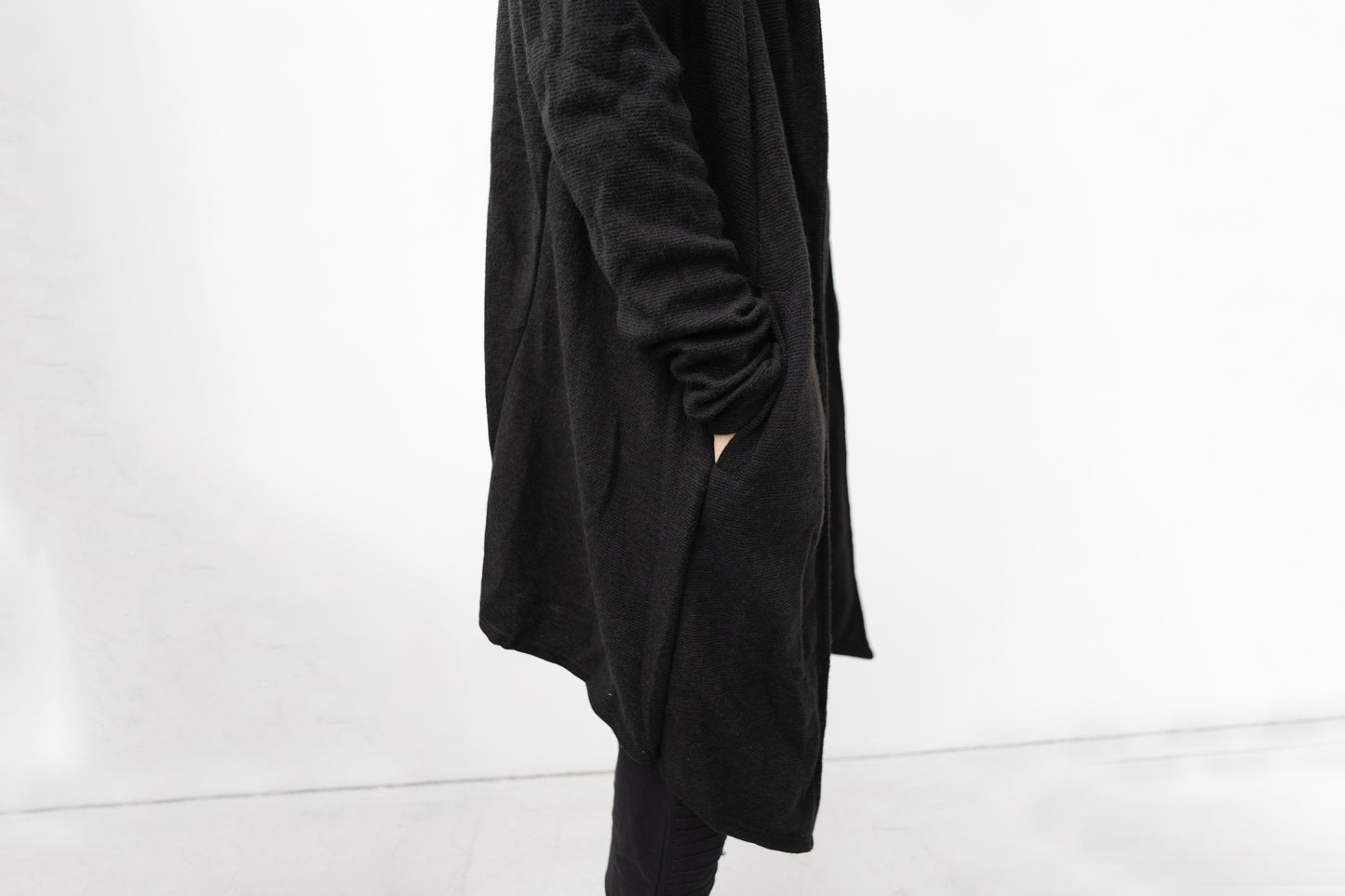 Detailansicht des schwarzen Strickmantels mit Kapuze, seitliche Tasche und strukturierte Oberfläche, Darkwear Mode mit klaren Linien.