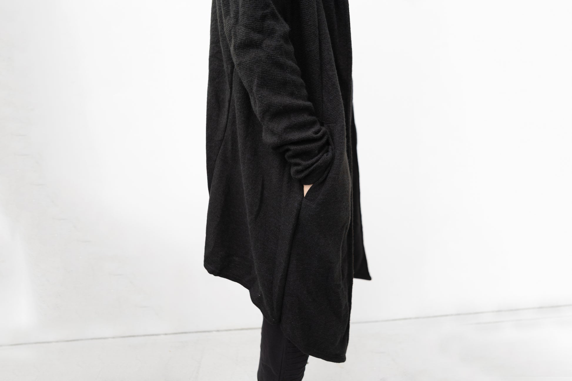 Detailansicht des schwarzen Strickmantels mit Kapuze, seitliche Tasche und strukturierte Oberfläche, Darkwear Mode mit klaren Linien.