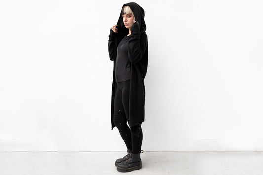 Model trägt langen schwarzen Strickmantel mit Kapuze im Goth-Stil, genderneutraler Schnitt, kombiniert mit schwarzen Leggings und Boots.