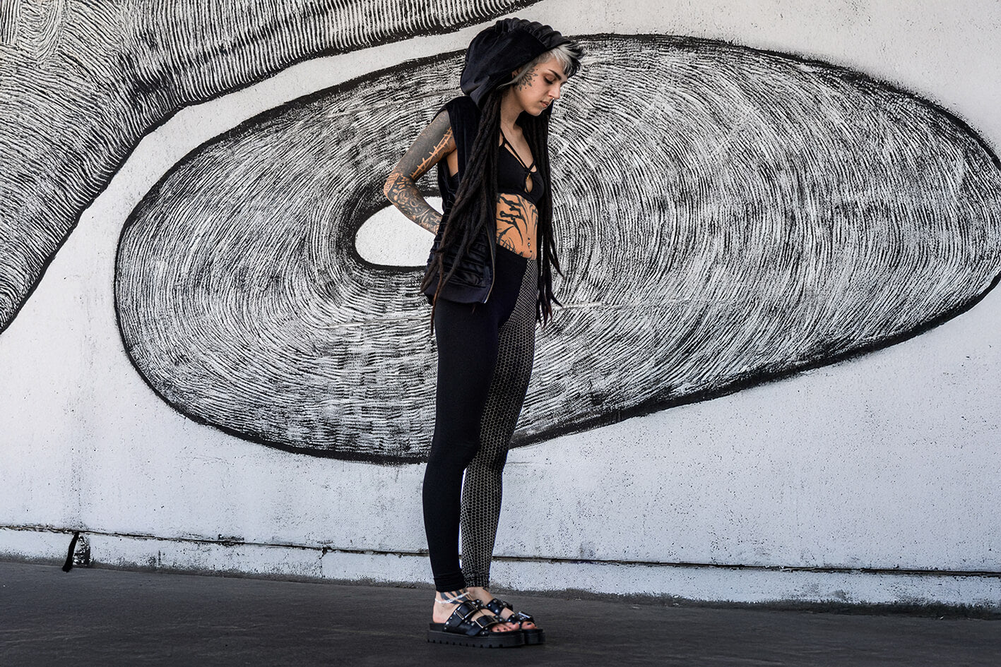 Stehendes Model vor Wand trägt Samtweste mit Kapuze, einen Strappy-Bra sowie Leggings mit abstraktem Ziegelstein-Print