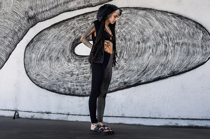 Stehendes Model vor Wand trägt Samtweste mit Kapuze, einen Strappy-Bra sowie Leggings mit abstraktem Ziegelstein-Print