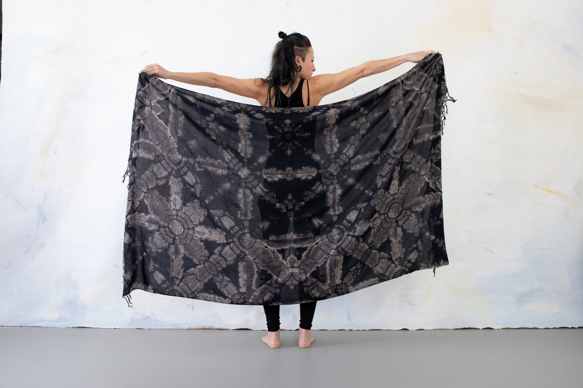 Model von hinten, das einen großen weichen Viskose-Sarong in Schwarz mit beige-grauen floralem Batikmuster und Fransen mit beiden Armen aufspannt